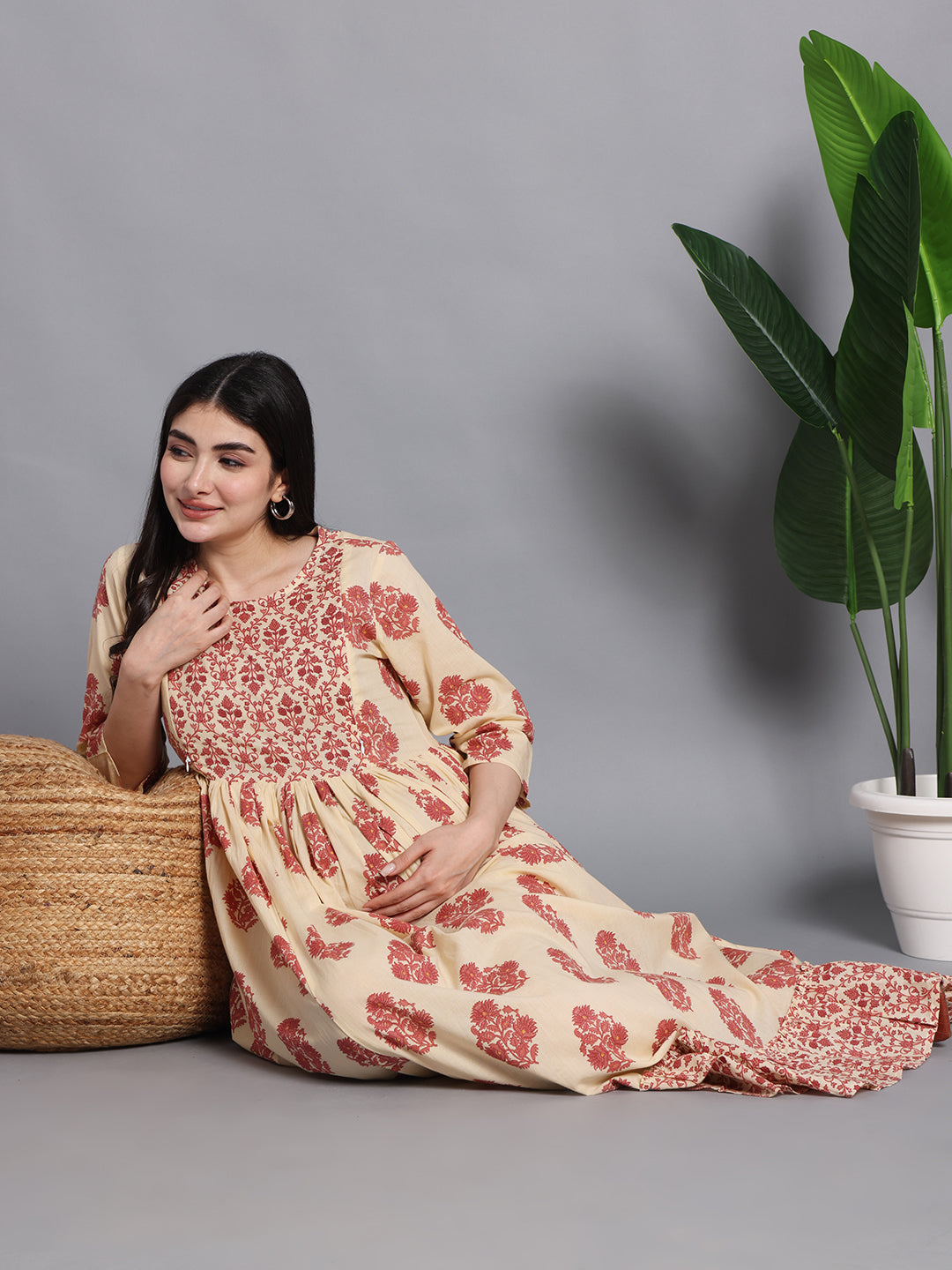 Pregnant woman in beige floral maternity dress sitting, indoor with a plant and woven ottoman
