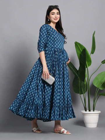 Pregnant woman in blue printed maternity dress, holding clutch, standing by potted plant