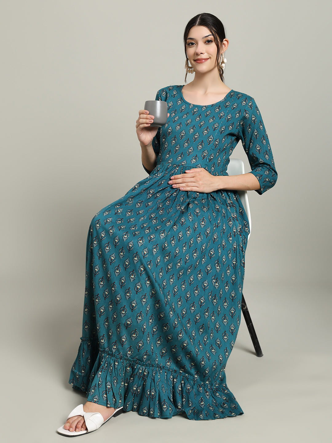 Smiling woman in teal printed maternity maxi dress, sitting and holding a cup