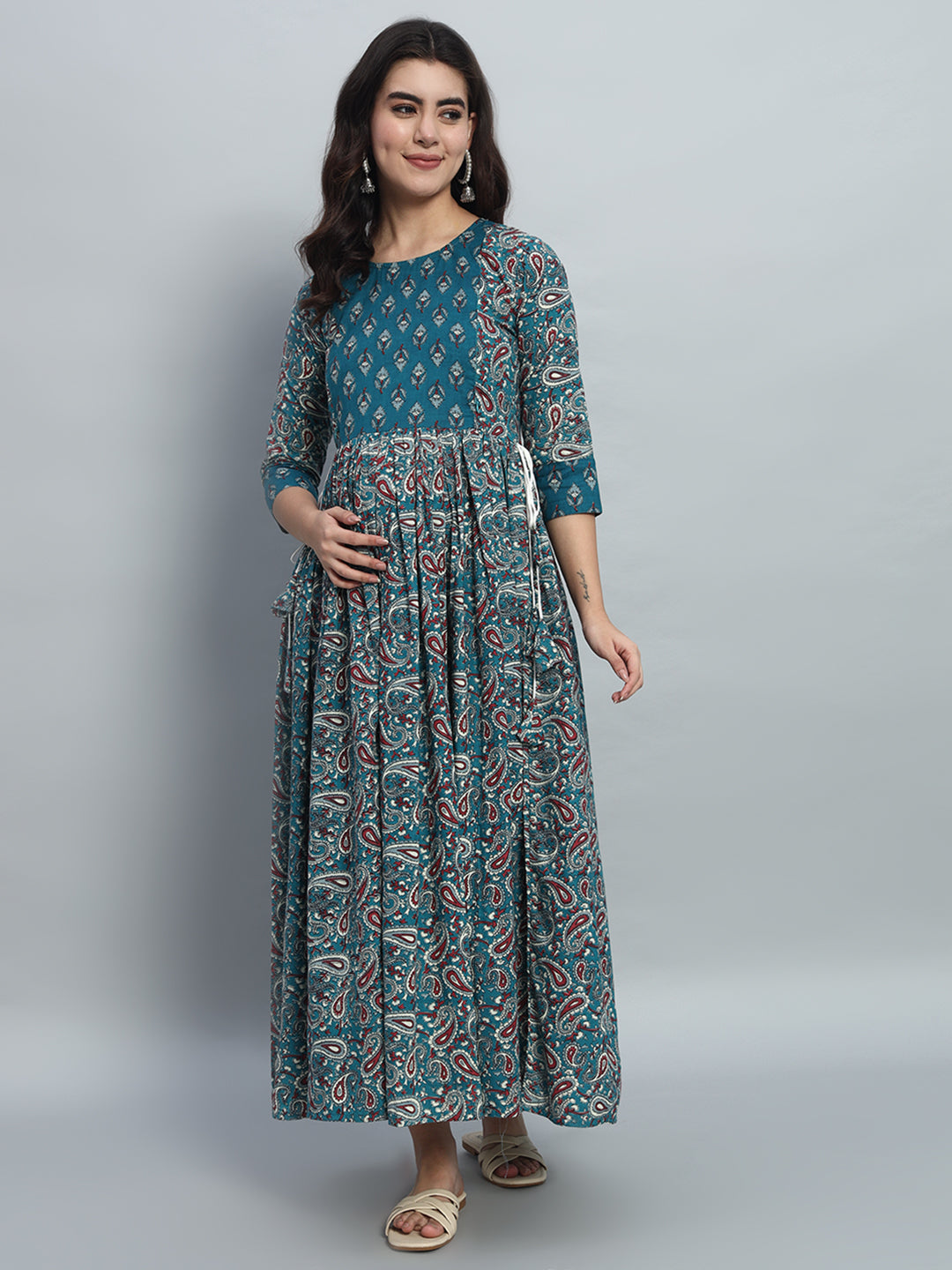 Pregnant woman wearing teal printed maternity maxi dress with three-quarter sleeves, posing indoors.