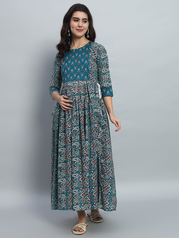 Pregnant woman wearing teal printed maternity maxi dress with three-quarter sleeves, posing indoors.