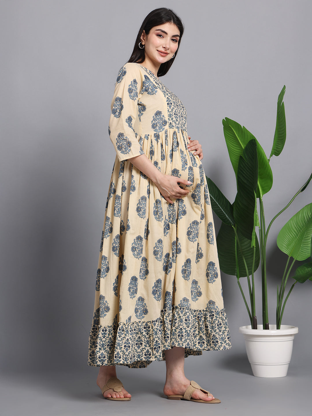 Pregnant woman in beige floral maternity dress posing indoors beside green potted plant