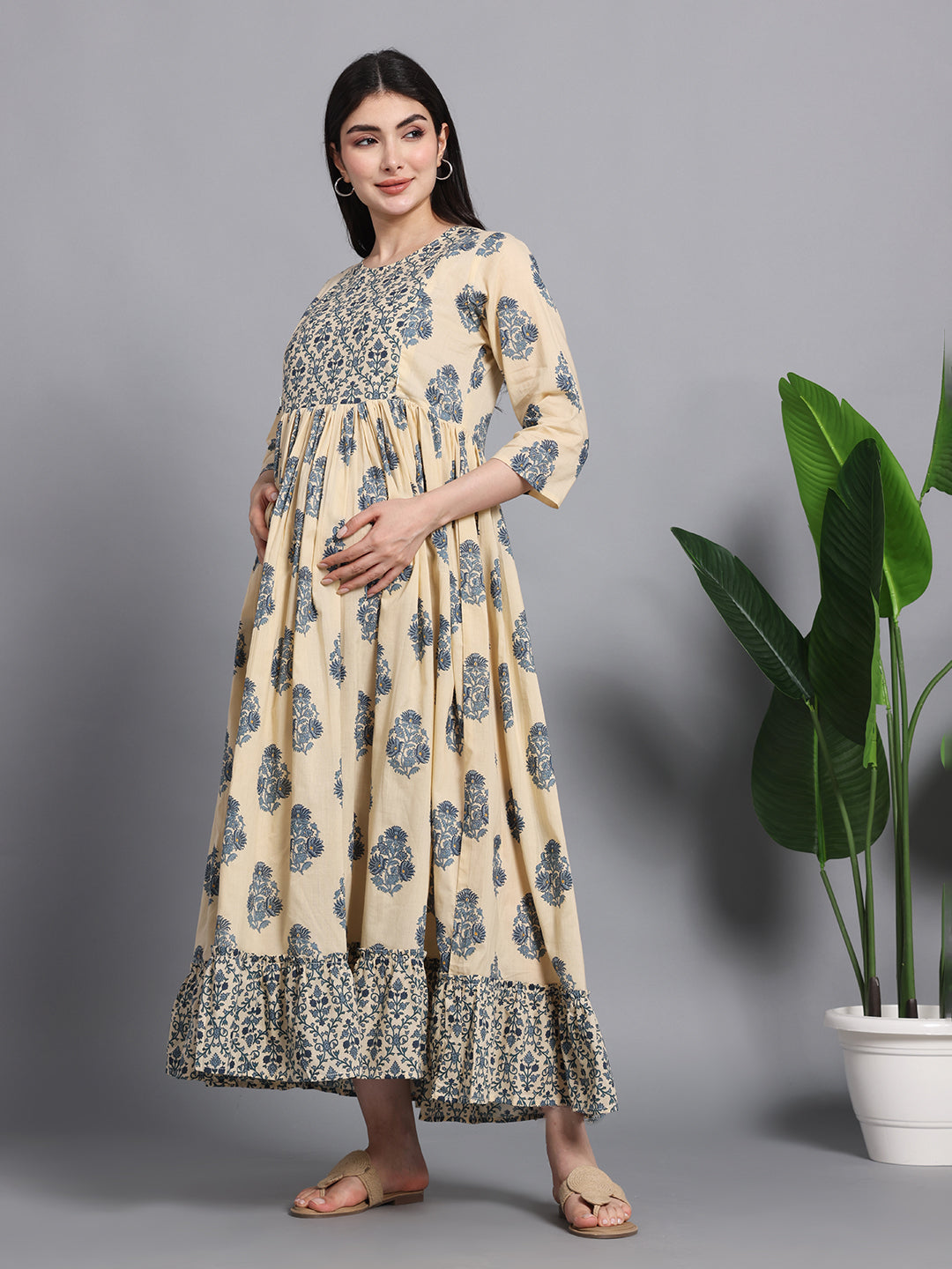 Pregnant woman wearing a beige and blue floral printed maternity maxi dress with three-quarter sleeves, standing indoors near a potted plant.