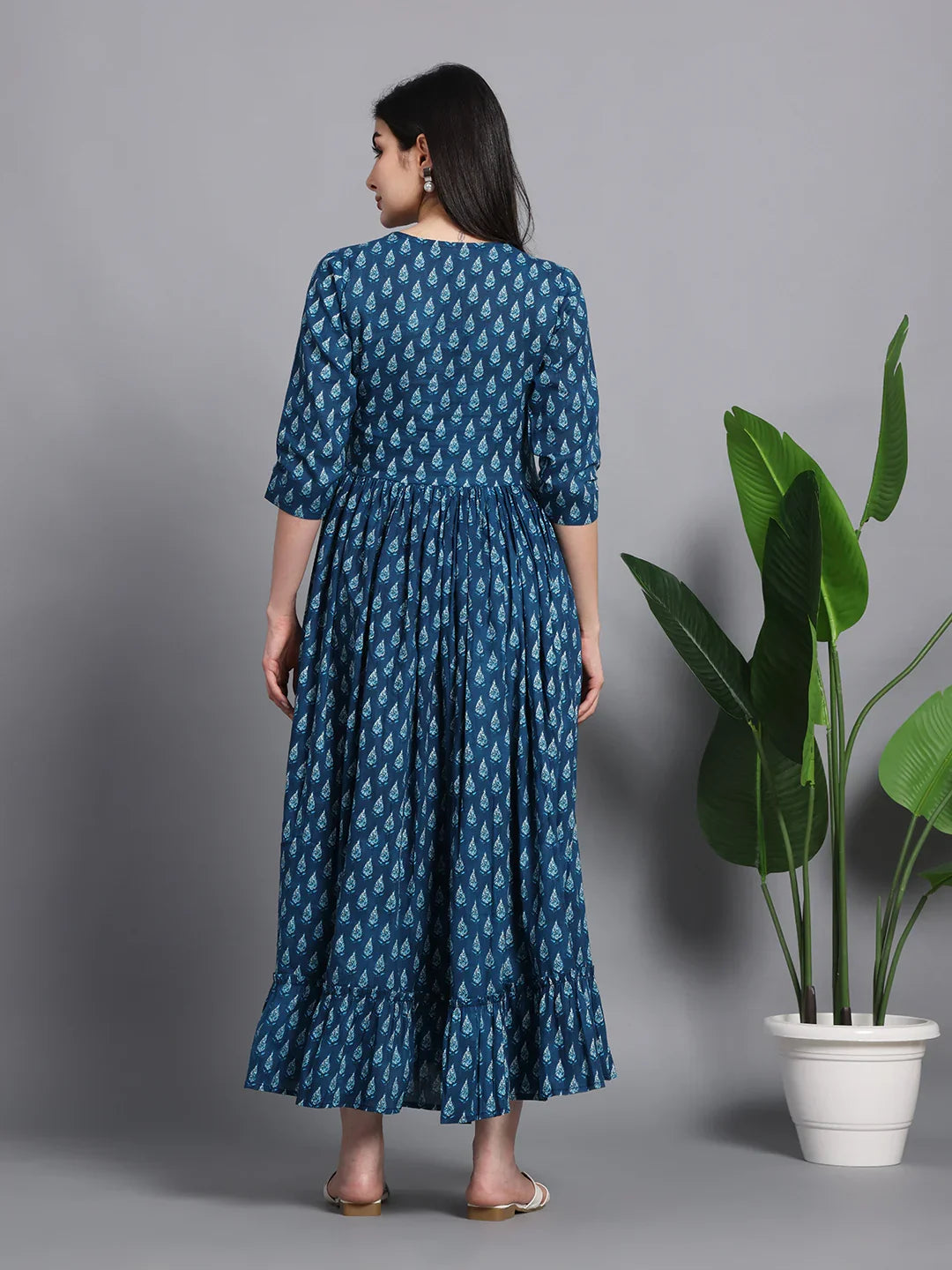 Woman in blue printed maternity maxi dress standing next to potted plant, back view