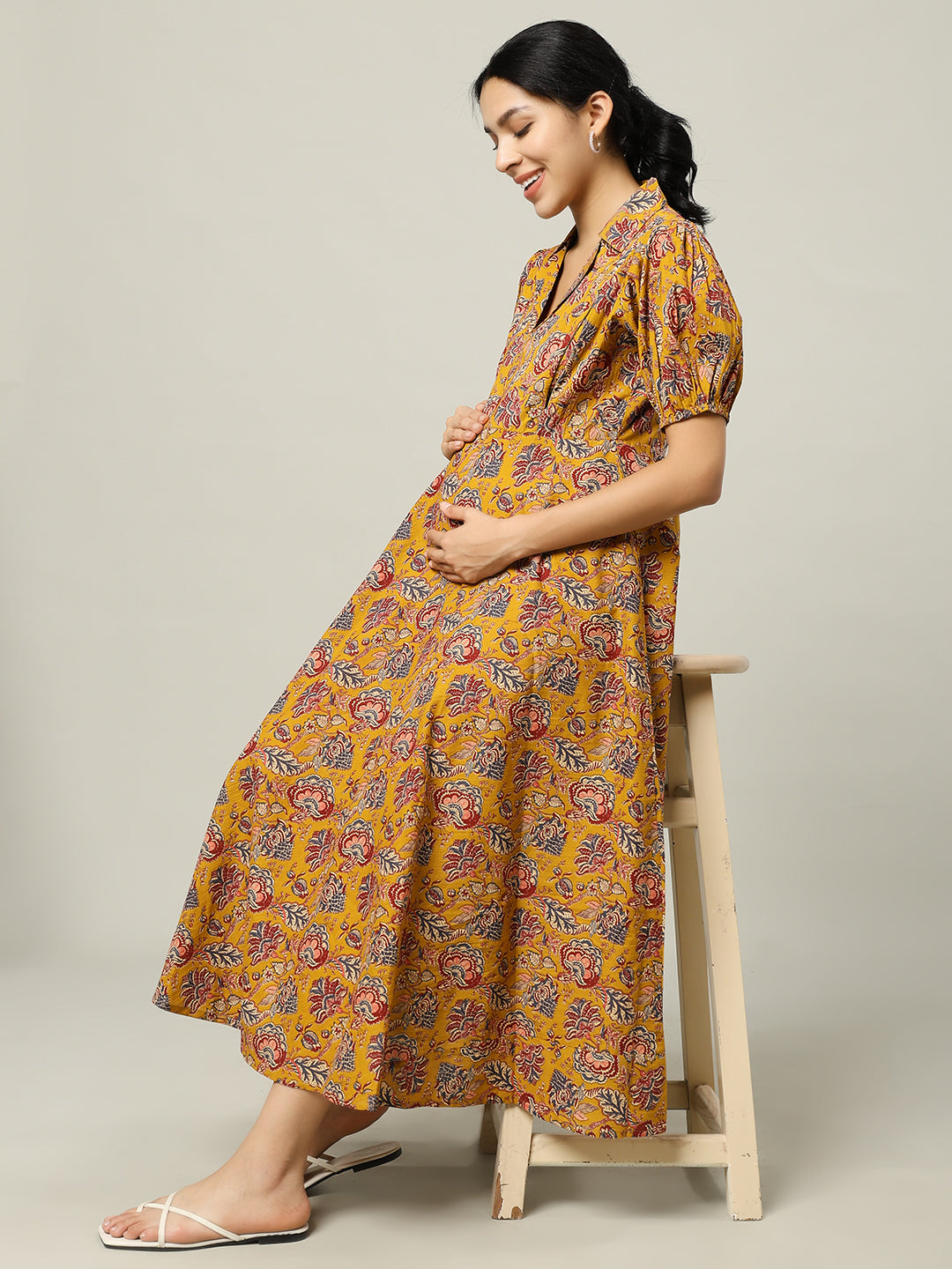 Pregnant woman in a yellow floral maternity dress sitting on a stool, smiling