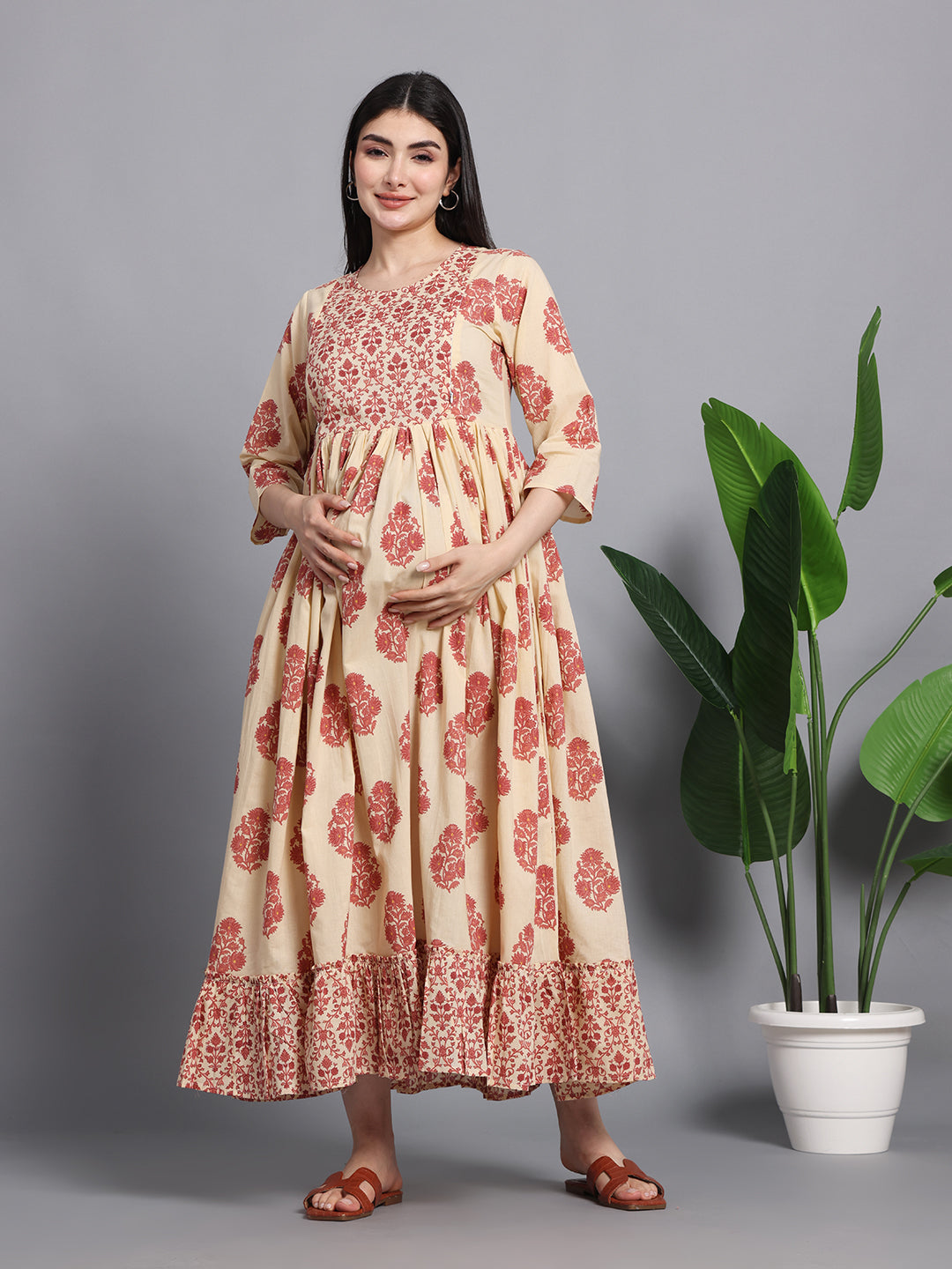 Pregnant woman in cream and red floral maternity dress with ruffled hem, standing indoors.