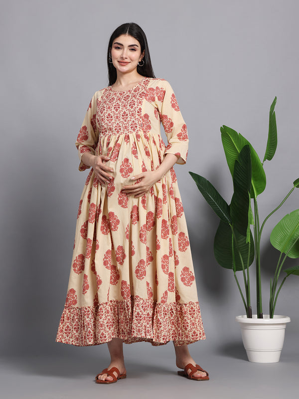 Pregnant woman in cream and red floral maternity dress with ruffled hem, standing indoors.