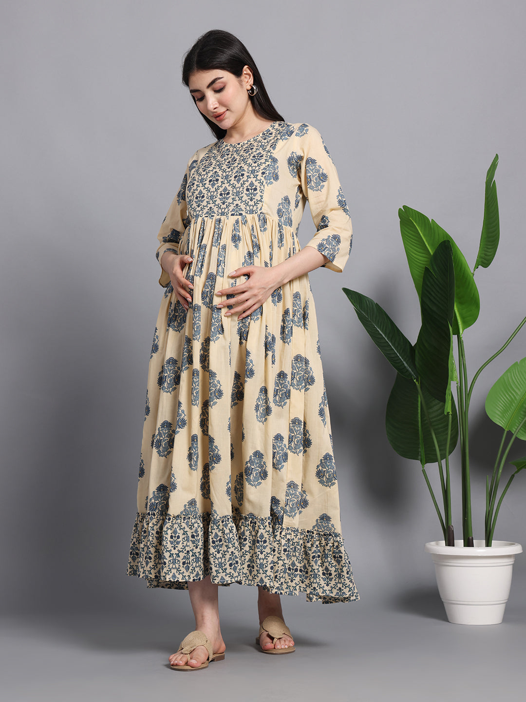 Pregnant woman in beige and blue floral maternity dress with three-quarter sleeves, standing indoors near green plant.