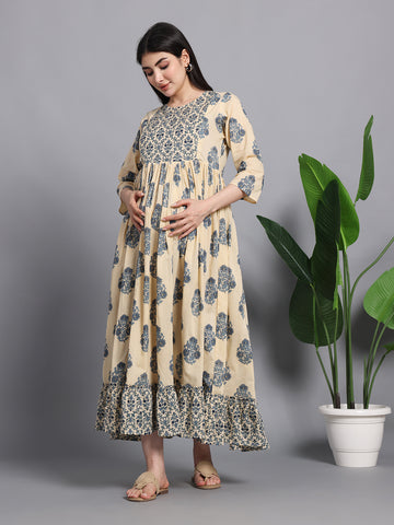 Pregnant woman in beige and blue floral maternity dress with three-quarter sleeves, standing indoors near green plant.