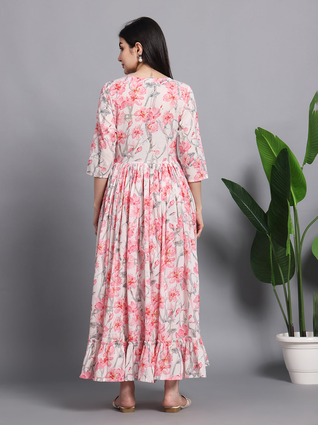 Woman wearing a pink floral maxi dress with ruffle hem, standing indoors near green plants