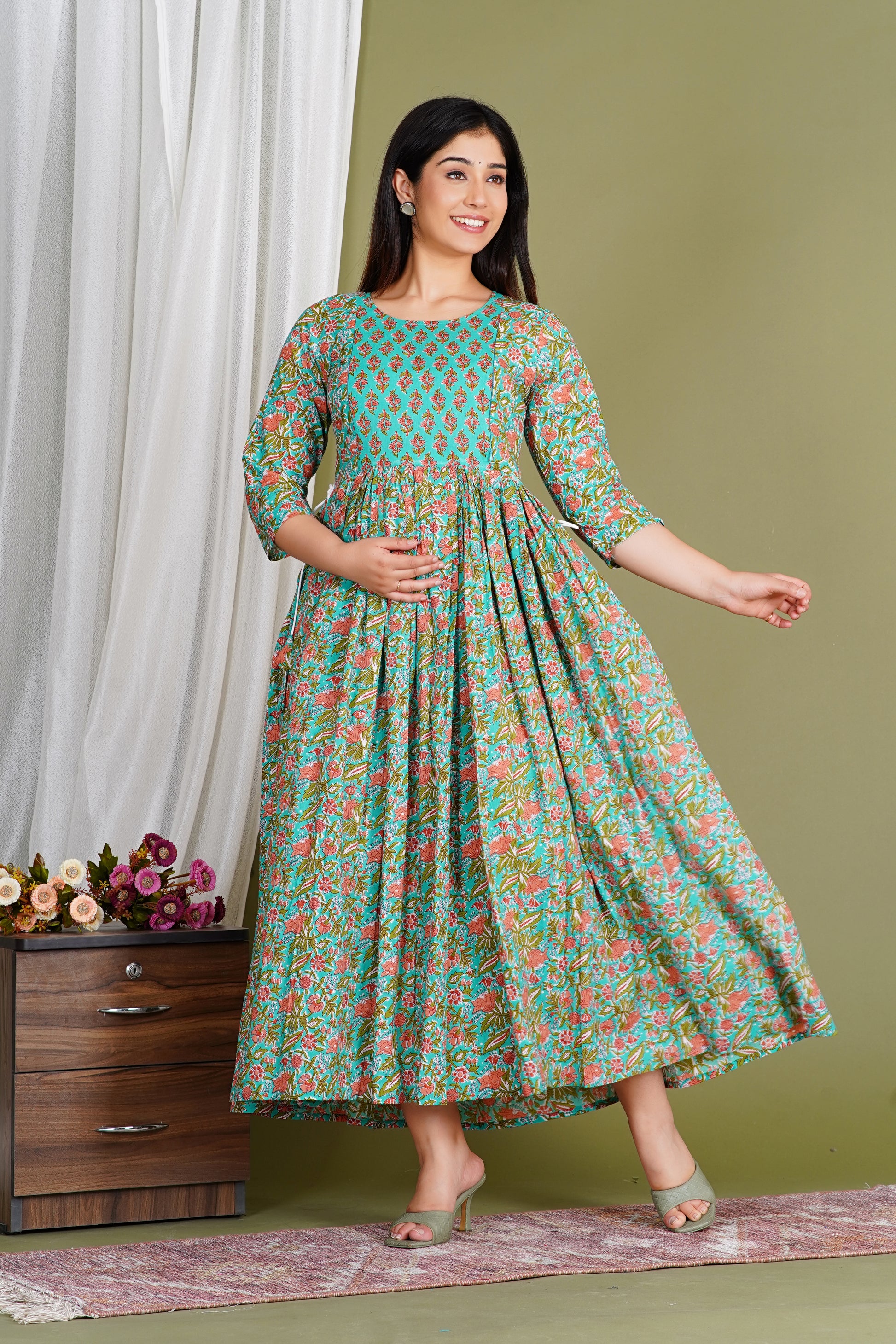 Smiling woman in a floral maternity dress with three-quarter sleeves, standing indoors.