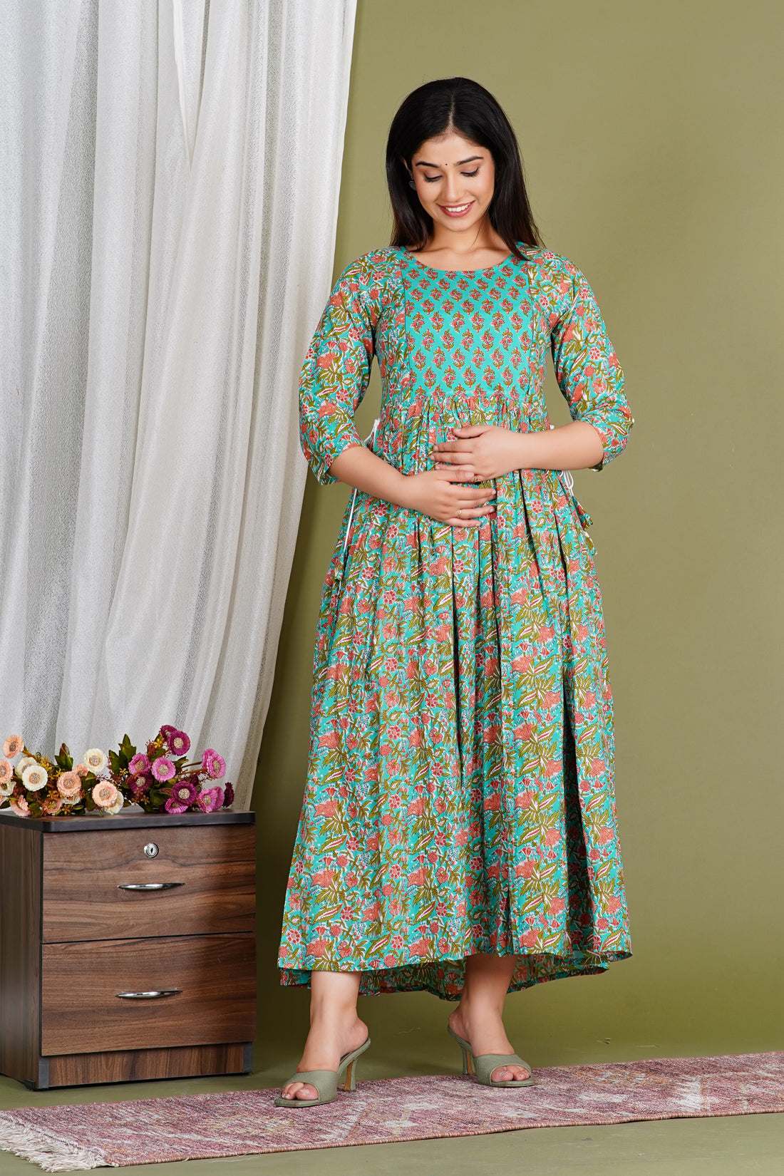 Smiling woman in a floral maternity dress with three-quarter sleeves, standing indoors.
