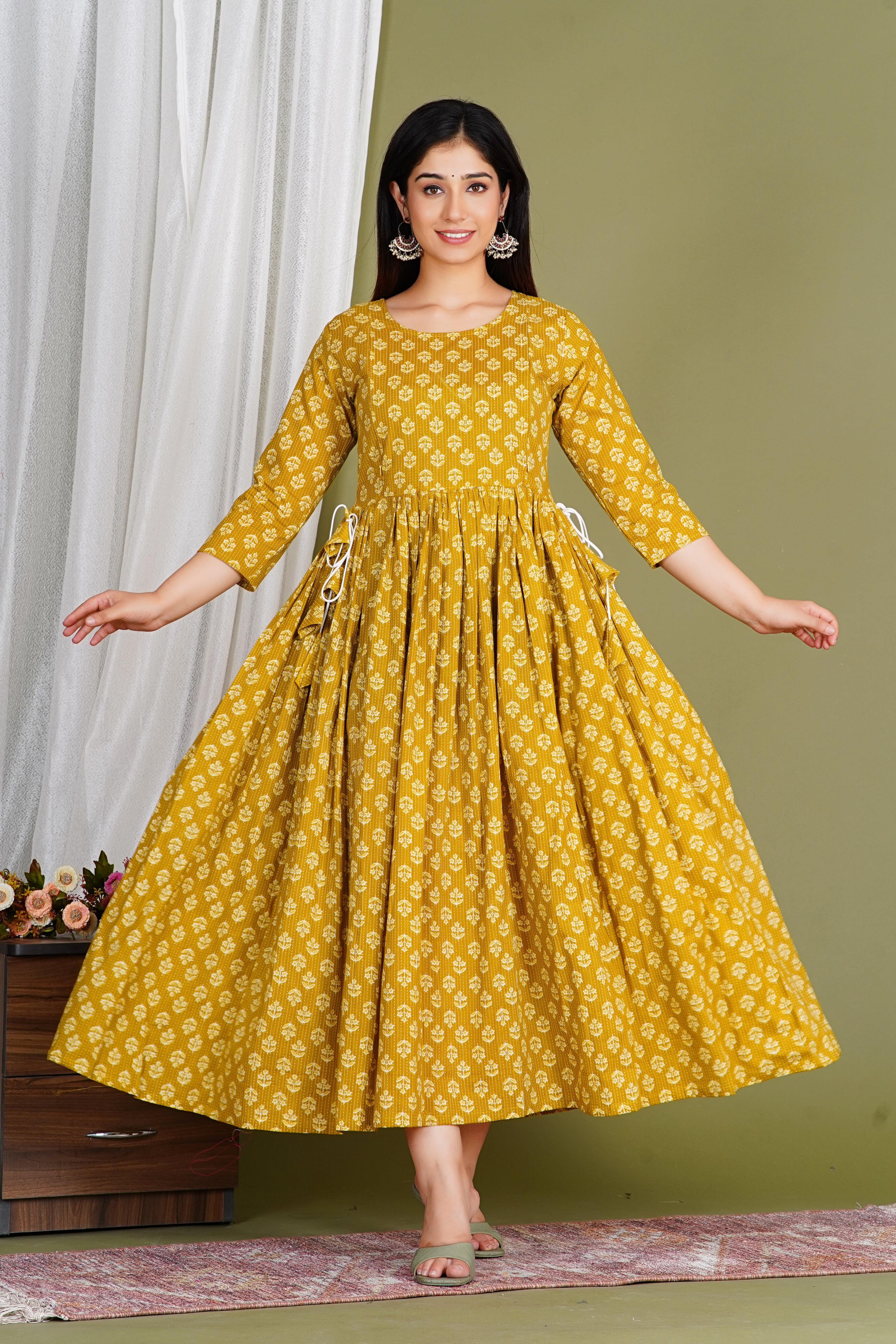 Woman in yellow floral maternity maxi dress with three-quarter sleeves and side ties, standing indoors.