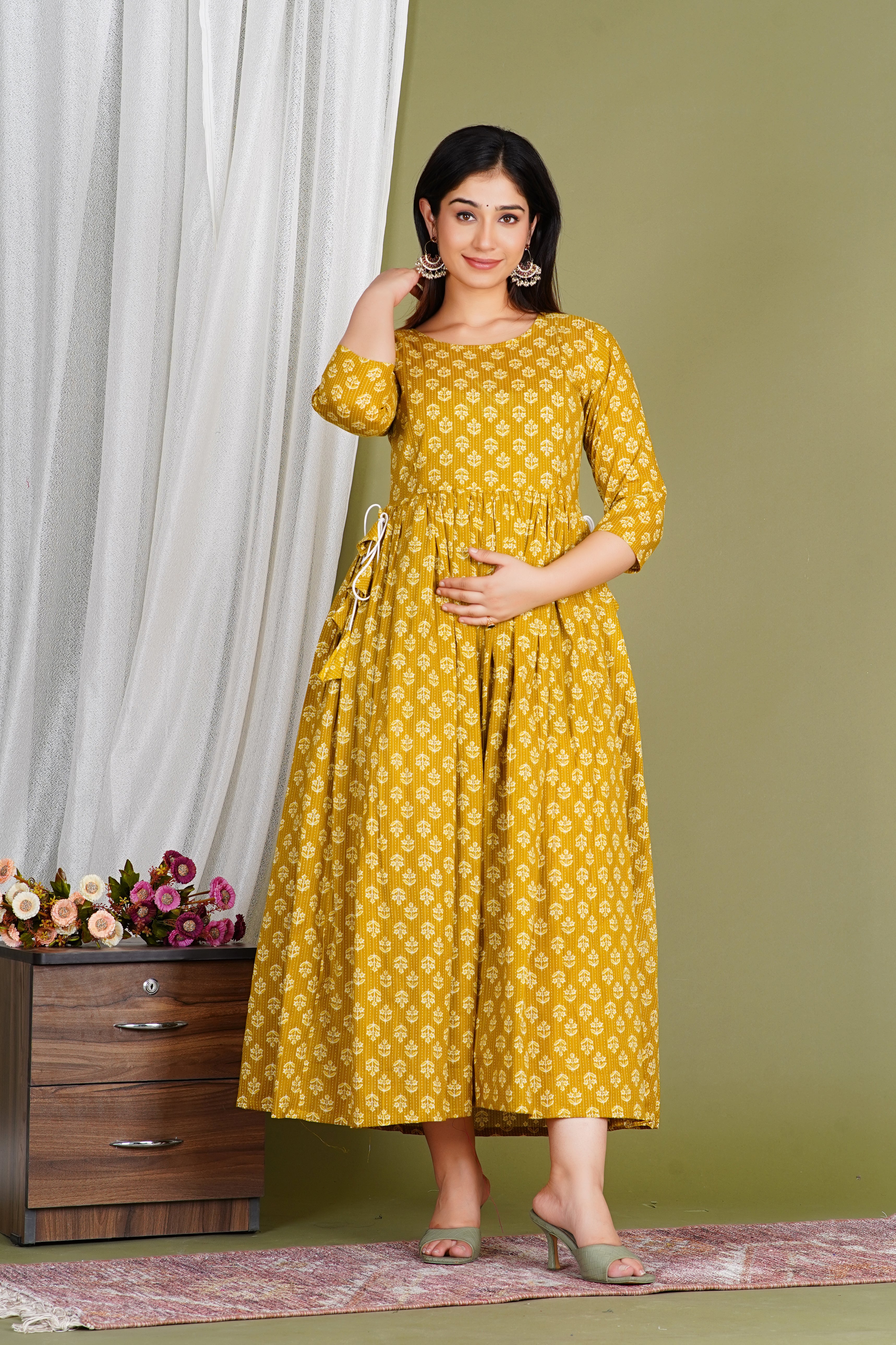 Woman in yellow printed maternity dress with side tie, posing indoors beside flowers